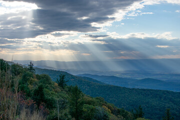 Fototapeta na wymiar Rays of light can be seen shining through the clouds over the blue Ridge Mountains inside Shenandoah National Park. The foreground of the photo features the brush and forests of a mountain.