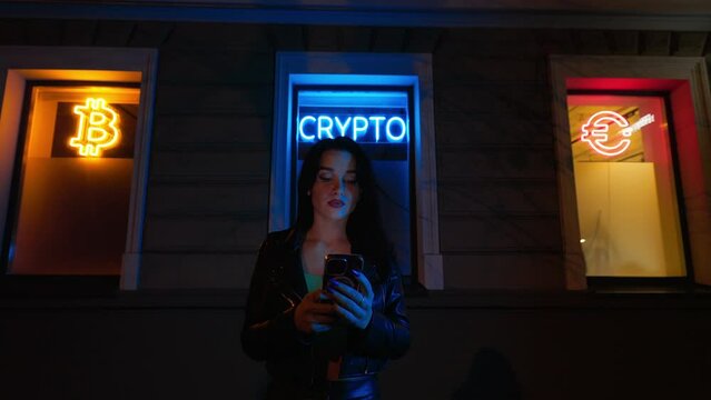 Young female crypto investor uses her smartphone buy or sell cryptocurrency at night against bright neon signs BITCOIN, CRYPTO and EURO on windows