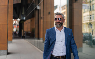 A happy hipster mature business man in glasses and a suit is walking through the city street near...