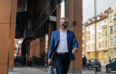 A handsome mature business man in glasses and a suit is walking through the city street near the office building.