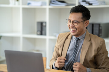 Excited happy Asian businessman looking at the computer laptop screen, celebrating an online win, overjoyed young asian male screaming with joy.