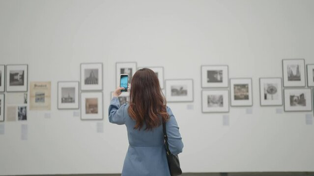 A young woman is taking photo of painting using camera of smartphone is photographing picture in gallery