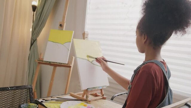 One young African American girl concentrates on acrylic color picture painting on canvas with a brush in art classroom, creative learning with talents and skills in elementary school studio education.