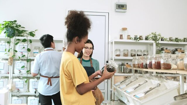 A female shopkeeper describes natural organic products to Black woman customer in refill store, zero-waste and plastic-free grocery, environment-friendly, sustainable lifestyles with a reusable shop.
