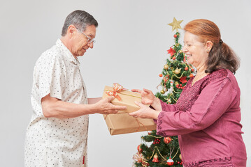 Senior latin couple smiling, spending the holidays at home, exchanging Christmas gifts near a decorated tree. The joy of celebrating together.