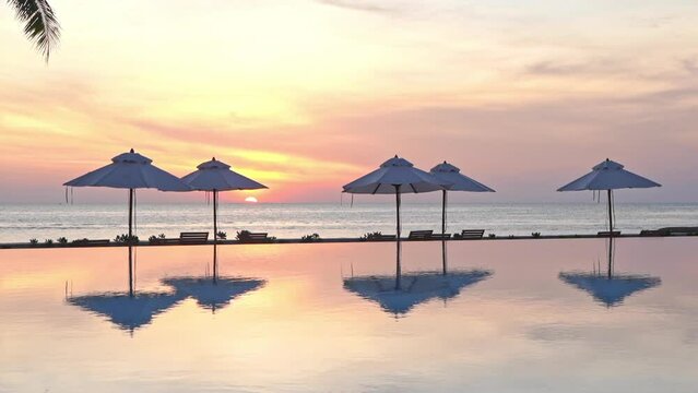 Colorful sunset along with beach umbrellas reflect in a resort swimming pool 