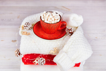A stack of knitted sweaters, a red cup of cocoa with marshmallows, a garland with Christmas decorations on a wooden background. Christmas winter composition, cozy home atmosphere. 