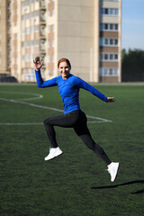 Women and sport. Smiling Girl in sportswear - blue shirt and black leggings does exercises: bouncing and having fun on the grass at the stadium outdoor on a sunny day. Middle aged sportswoman