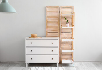 Shelving unit with flowerpot, chest of drawers and folding screen near light wall