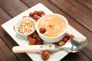 Plate with bowls of tasty nut butter and hazelnuts on wooden background, closeup