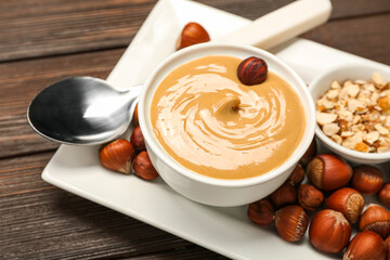 Plate with bowl of tasty hazelnut butter and nuts on wooden table, closeup