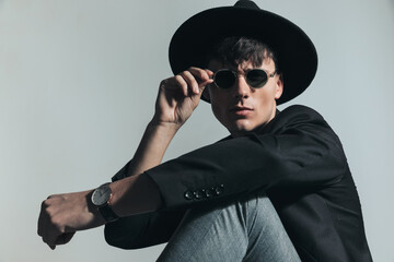 stylish young man with hat holding elbow on knee and fixing sunglasses