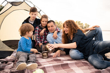 Happy family resting in a tent in nature. Summer vacation with children. Large family with three children drinking tea outdoors at the campsite