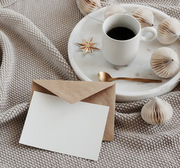 Obraz na płótnie Canvas Blank greeting card, invitation mock up, craft envelope,beige knitting blanket, Christmas tree decorations in marble tray and cup coffee.Xmas scene stationery still life neutral colors.Copy space