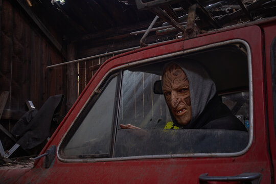 The Monster Looks Out Of The Window Of An Old Red Car. A Man In A Halloween Mask Was About To Leave An Old Wooden Cluttered Barn. Dark Photo. Selective Focus.