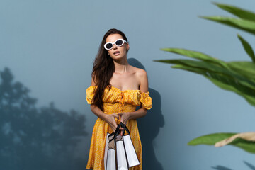 Pretty woman in bright yellow summer dress posing on blue wall background wearing trendy sunglasses holding stylish shopping bags happy excited, sale black friday