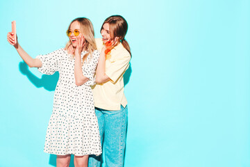 Two young beautiful smiling brunette hipster female in trendy summer clothes. Sexy carefree women posing near blue wall. Positive models having fun. Cheerful and happy