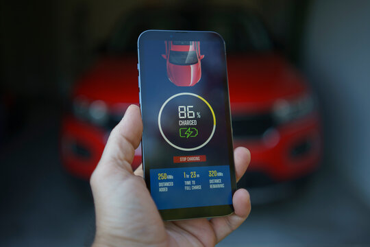 Using mobile smartphone device refueling electric vehicle EV car mock UI application display screen, hand holding cellphone at electric car charging station connected power cable alternative energy