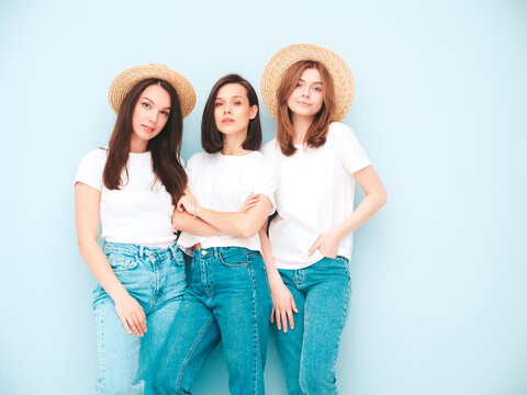 Three young beautiful smiling hipster female in trendy same summer white t-shirt and jeans clothes. carefree women posing near light blue wall in studio. Cheerful positive models having fun. In hats