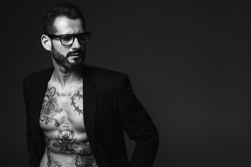 Fototapeta na wymiar Portrait of handsome confident stylish hipster lambersexual model. Sexy modern man dressed in black jacket. Naked torso with tattoos.Fashion male posing in studio on dark background. In spectacles