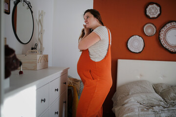 Pregnant plus size middle-aged woman combing her hair in front of mirror in bedroom.
