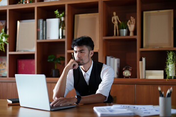 A portrait of a good-looking and discreet Asian man sitting at his desk with a thoughtful look on his computer