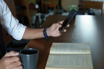 A portrait of a handsome and discreet Asian man sitting at his desk using his phone while taking a break from reading and drinking coffee