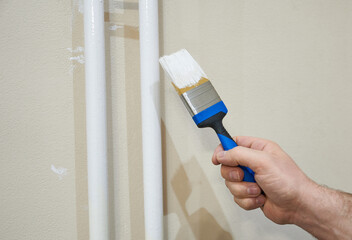 A man's hand with a brush in white paint against a background of painted pipes. The concept is an apartment renovation.