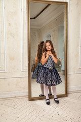 Little girl princess 5-6 years old, thoughtful looking at camera, fashion model in stylish elegant blue dress stands in living room with mirror. Fashionable young lady actress at home. Copy text space