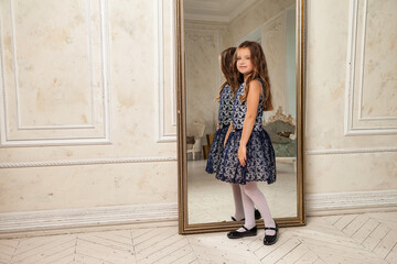Princess little girl 5-6 years old, thoughtful looking at camera, fashion model in stylish elegant blue dress standing in living room with mirror. Fashionable young lady actress at home. Copy space