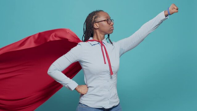 African american superhero flying with red cape and costume over blue background. Wearing cloak with hero costume to pose as action cartoon character, showing victory and courage.