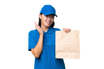 Young Asian woman taking a bag of takeaway food over isolated background saluting with hand with happy expression