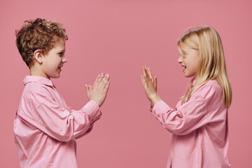 happy children, a boy and a girl, in pink clothes stand sideways to the camera and play clapping their hands with each other standing on a pink background