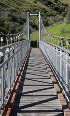 Fototapeta premium Suspension metal bridge across the river with a green hill in the background, interesting geometric picture, vertical view
