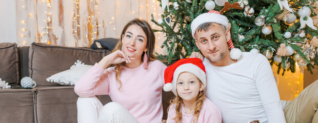 Christmas family. Happy portrait dad, mom and daughter in santa hats sitting on the floor in the living room at home near the christmas tree, all smiling with festive mood from winter holidays