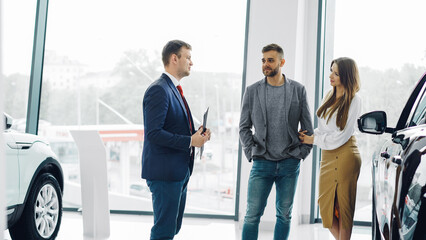 Professional car salesman is telling interested buyers beautiful couple about luxurious car in motor show while man and woman are looking at auto and listening to polite dealer.