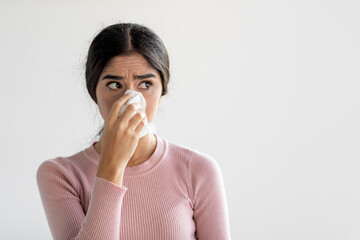 Serious young indian lady sneezes in napkin and blows her nose on gray wall background