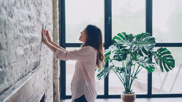 Creative young woman is decorating her loft style apartment choosing place on brick wall for beautiful picture and marking spot with pencil. Creativity and interior concept.