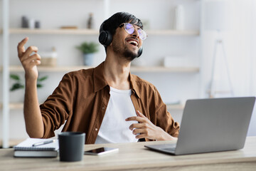 Happy indian guy with headset sitting at workdesk, singing