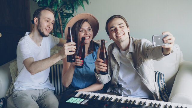 Happy attractive musicians are taking selfie with beer bottles sitting on sofa in home studio and holding smartphone. Young people are posing, clanging bottles and having fun.