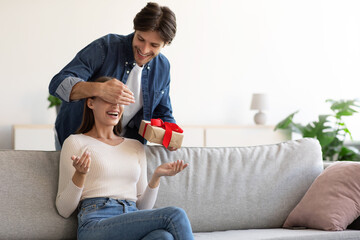 Smiling millennial european guy closes eyes to woman, gives present in box to surprise smiling wife