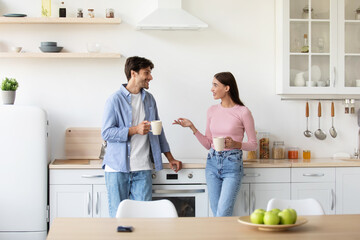 Cheerful smiling young wife and husband with cups of hot drink talk on kitchen interior in morning