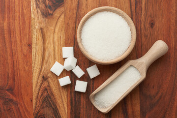 Obraz premium Miseczka i łopatka z białym cukrem, obok duże kostki białego cukru na drewnianym tle