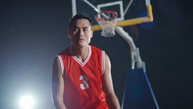 Basketball player hits the ball on the floor, warms up before the game, asian basketball player looks at the camera, indoor playground.