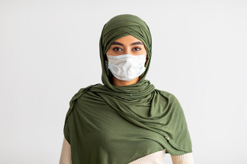 Pretty millennial Arab female in hijab wearing medical mask during coronavirus epidemic on light studio background