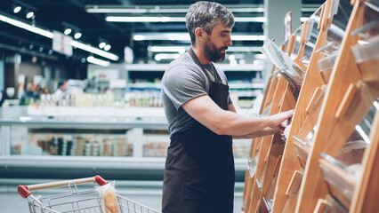 Busy male salesman is putting bread on shelves in bakery department in food store, bearded guy is...