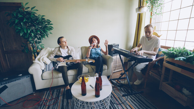 Musical band is rehearsing at home, blond woman is singing and men are playing musical instruments guitar and keyboard. Beer bottles and snacks on modern table are visible.