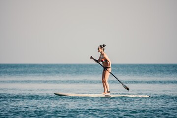 Sea woman sup. Silhouette of happy middle aged woman in rainbow bikini, surfing on SUP board, confident paddling through water surface. Idyllic sunset. Active lifestyle at sea or river.