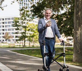 Smiling senior man using mobile phone listening to music through wireless headphones walking with mobility walker on footpath