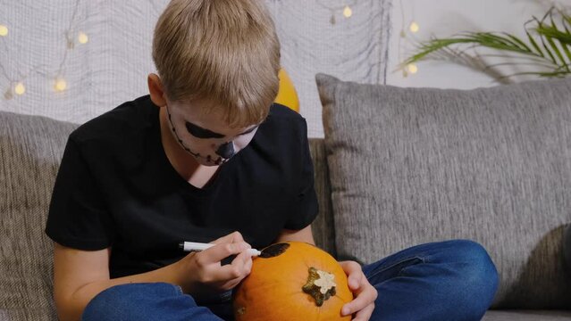 A boy with a painted skeleton on his face paints a pumpkin for Halloween.